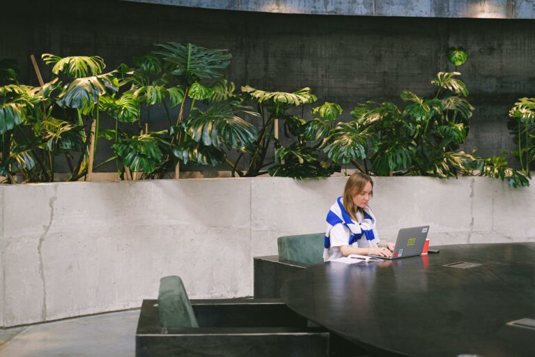 Woman working on a laptop in a modern indoor setting with lush greenery and sleek furniture.